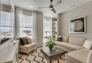 Modern living room with beige sectional sofa, large windows, and a ceiling fan. Decorative shelf and plant in view.